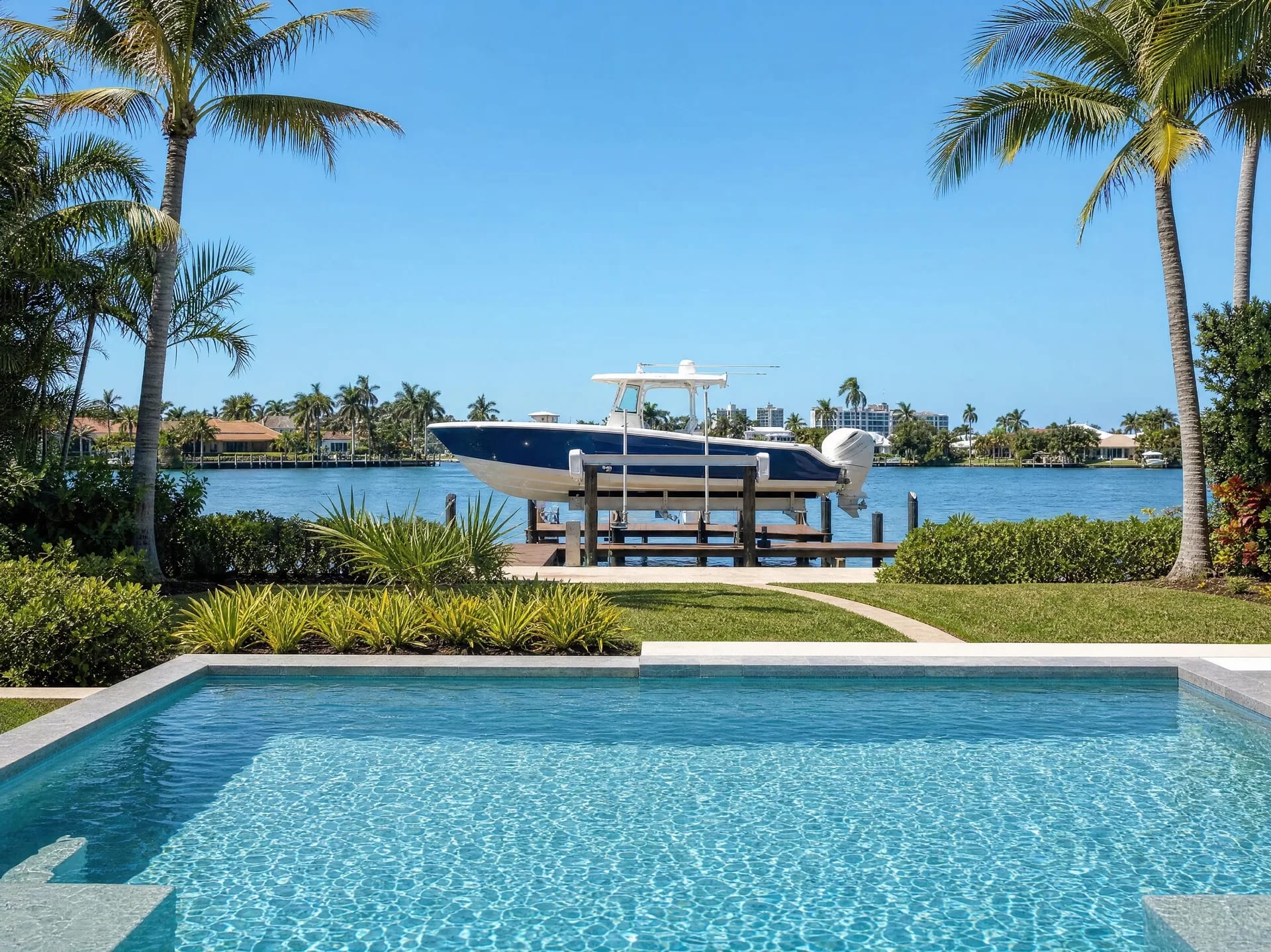 A center-console boat on its lift at a private waterfront dock, with a pool and palm trees in the foreground